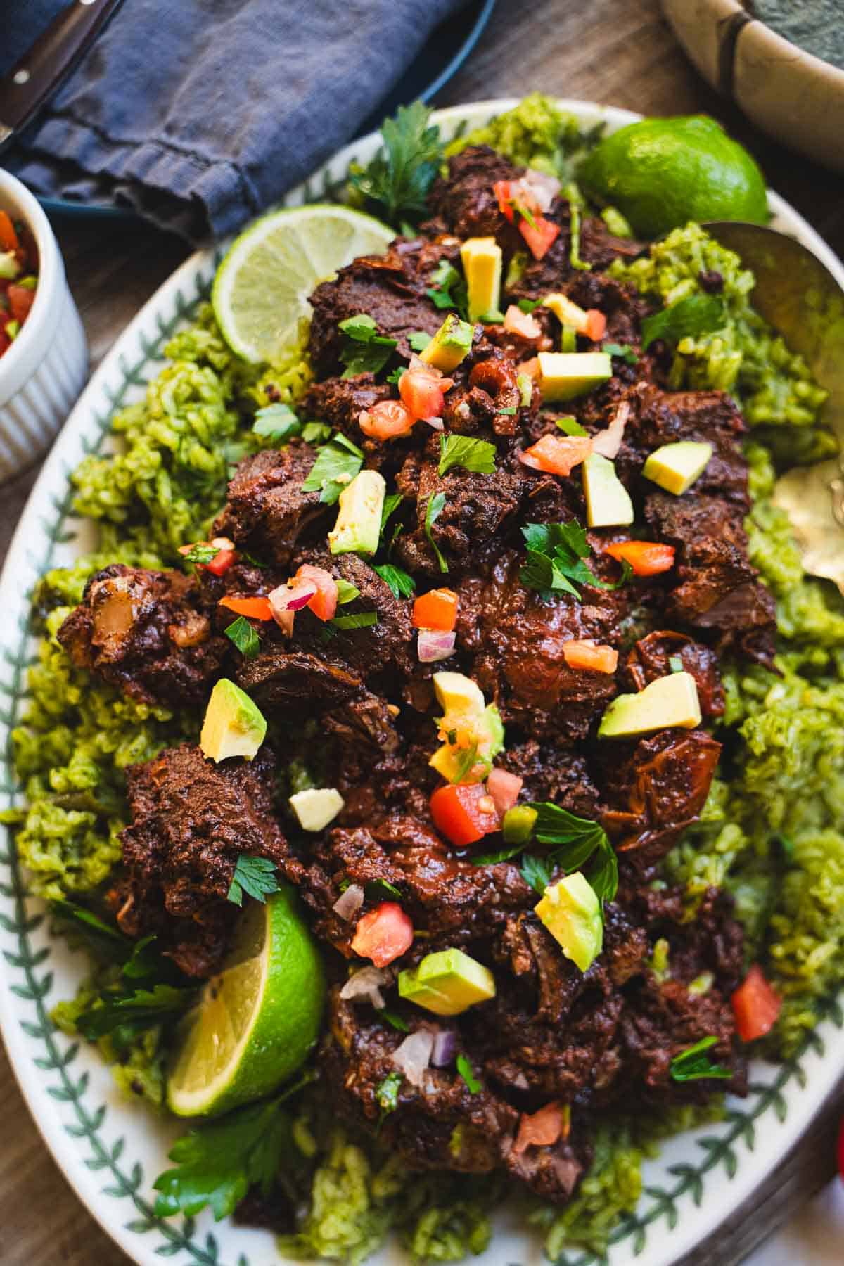 Lamb barbacoa on a white platter over a bed of mexican green rice garnished with tomatoes, feta, lime slices.