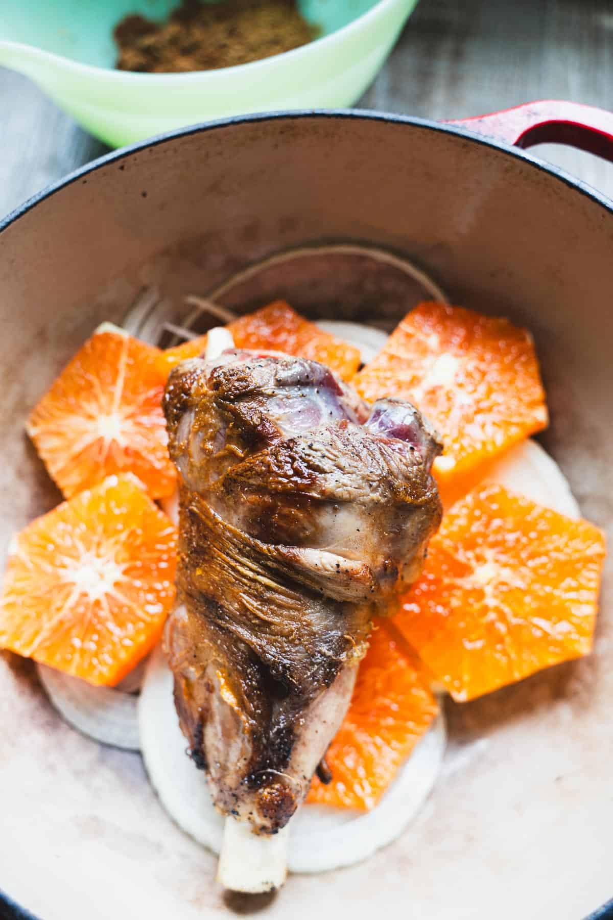 Orange slices without rind and sliced onion nestled around lamb shank in a dutch oven.