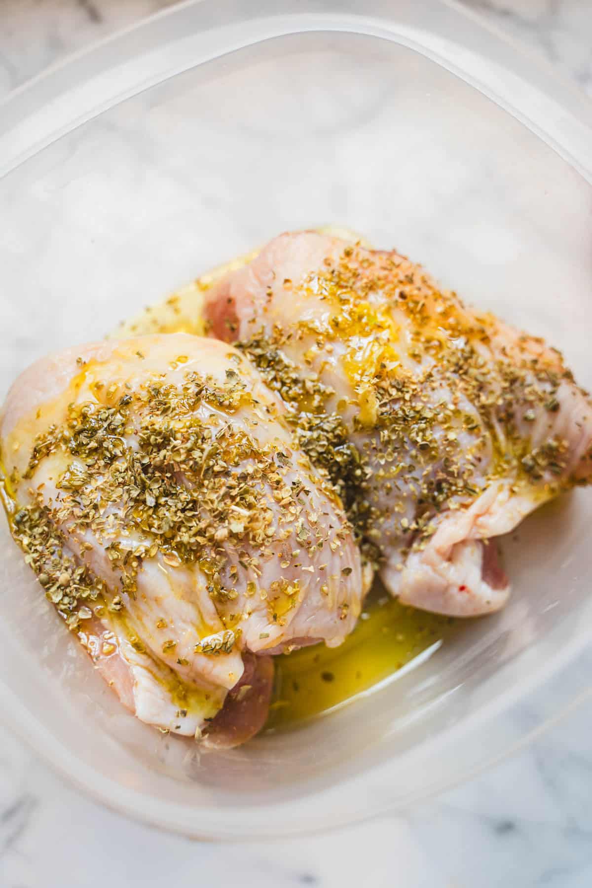 Skin on, bone-in chicken thighs in a dried oregano and olive oil marinade in a plastic container.