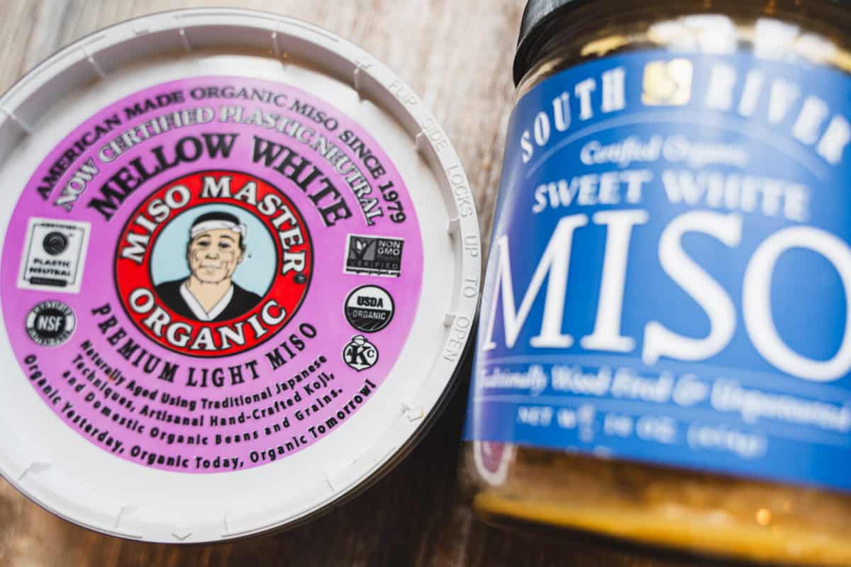 Two brands of miso one miso paste in a white container and another white miso in a jar.