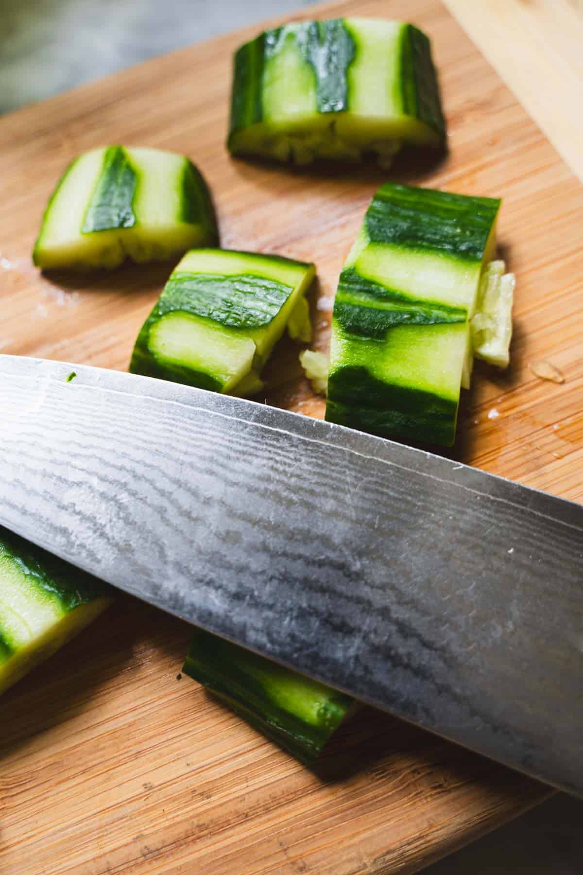 Flat edge of knife smashing cucumber slices on a wooden cutting board to make smashed cucumber salad.