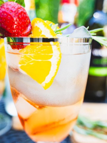 Rose port and tonic in a balloon wine glass with ice and a garnish of rosemary, orange and strawberry on resting over the lip of the glass.