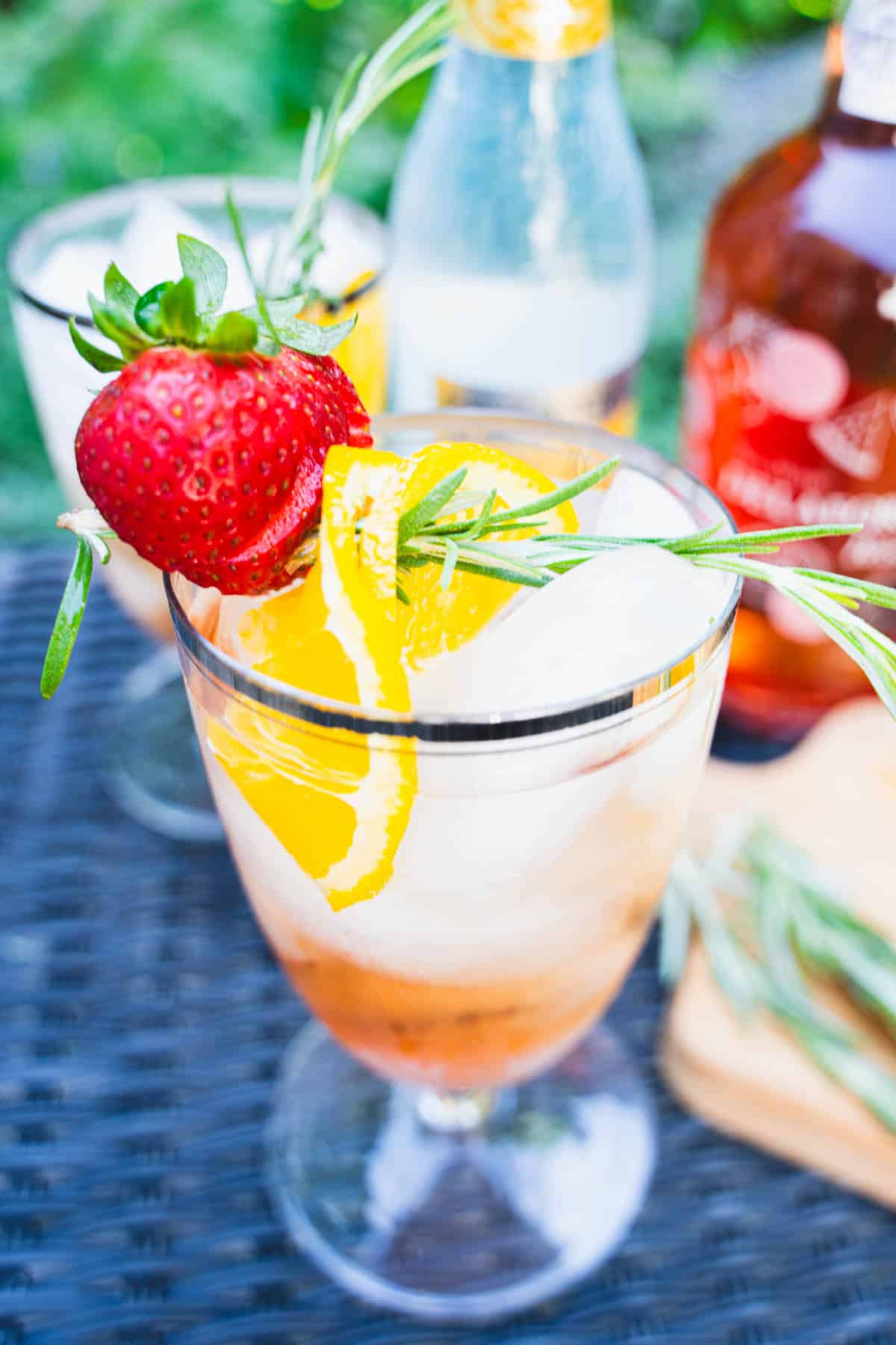 A thin slice of orange and a strawberry thread on a rosemary stick resting on top of the lip of the cocktail glass.
