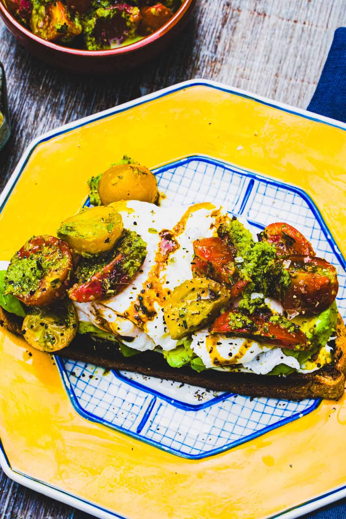 Avocado toast with marinated tomatoes and poached egg on yellow and blue plate next to a bowl of marinated tomatoes and a salt jar.