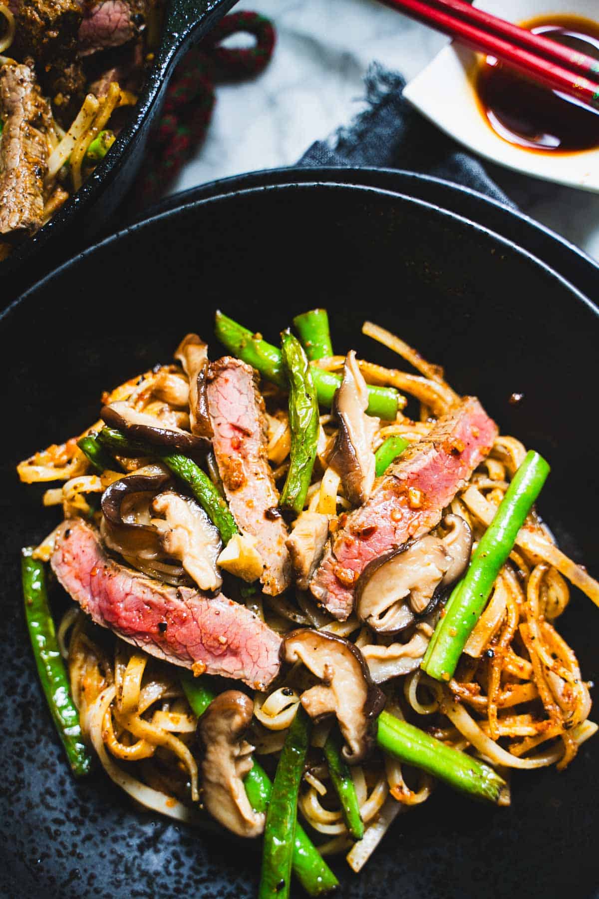 pan-fried green beans and rice noodles tossed in a chili crisp sauce garnished with pan seared flank steak and pickled mushrooms in a black bowl with chili crisp in a white bowl next to the the beef and noodles in the black bowl.