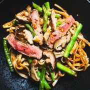 pan-fried green beans and rice noodles tossed in a chili crisp sauce garnished with pan seared flank steak and pickled mushrooms in a black bowl with chili crisp in a white bowl next to the the beef and noodles in the black bowl.
