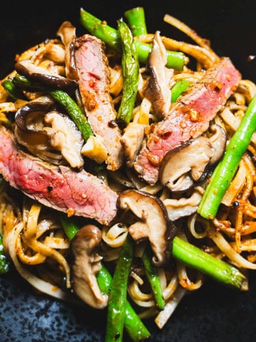pan-fried green beans and rice noodles tossed in a chili crisp sauce garnished with pan seared flank steak and pickled mushrooms in a black bowl with chili crisp in a white bowl next to the the beef and noodles in the black bowl.
