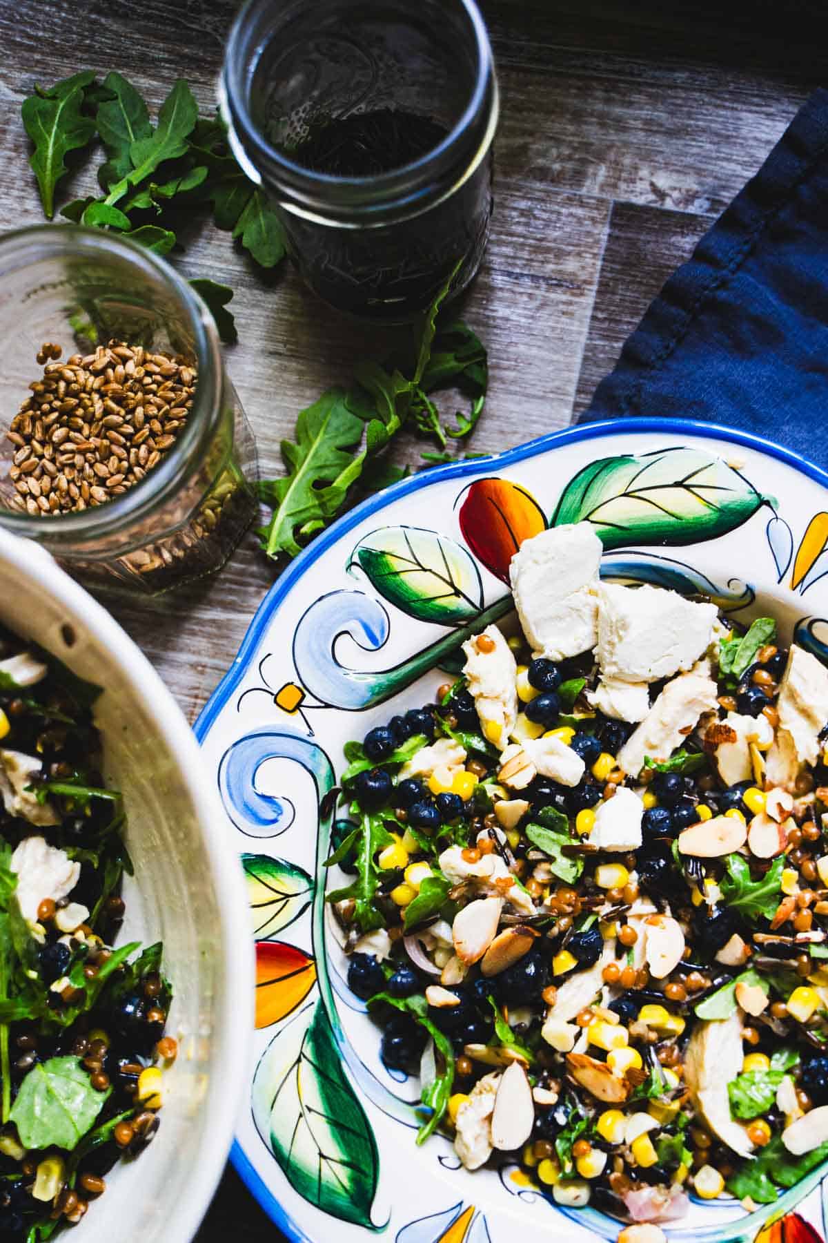 Wheat berry and wild rice grain bowl with pickled corn and blueberries in a decorated bowl and a salad bowl with wheat berries and wild rice in glass jars.