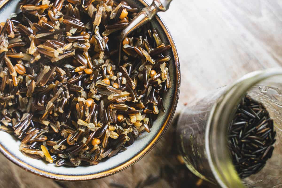 Cooked wild rice in a bowl with a spoon and uncooked wild rice in a glass jar.