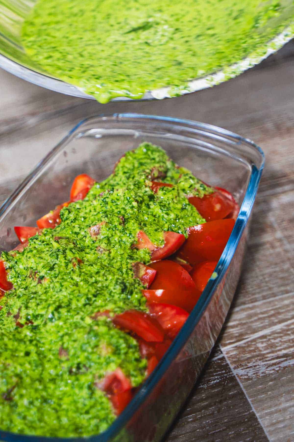 Pureed fresh herb marinade being poured over quartered cherry tomatoes in a glass dish.