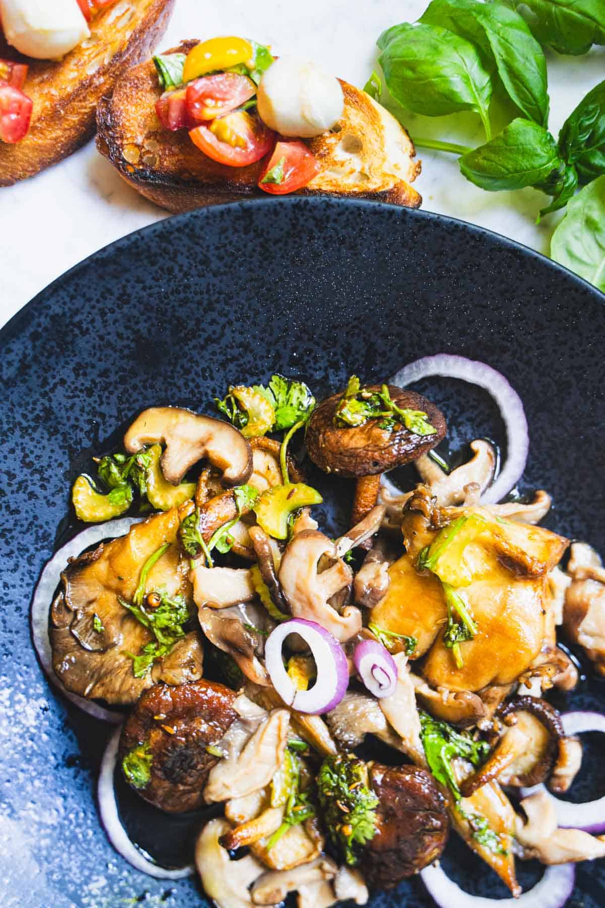 Mushroom salad tossed with pickled mushrooms, roasted mushrooms, celery, cilantro, red onions and a vinaigrette on a black plate next to cherry tomato bruschetta and sprigs of basil.