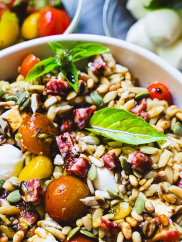 Orzo salad in a bowl with marinated cherry tomatoes, roasted eggplant cubes, soppressata cubed and basil. Alongside a bowl of marinated cherry tomatoes and a bowl of mozzarella balls.