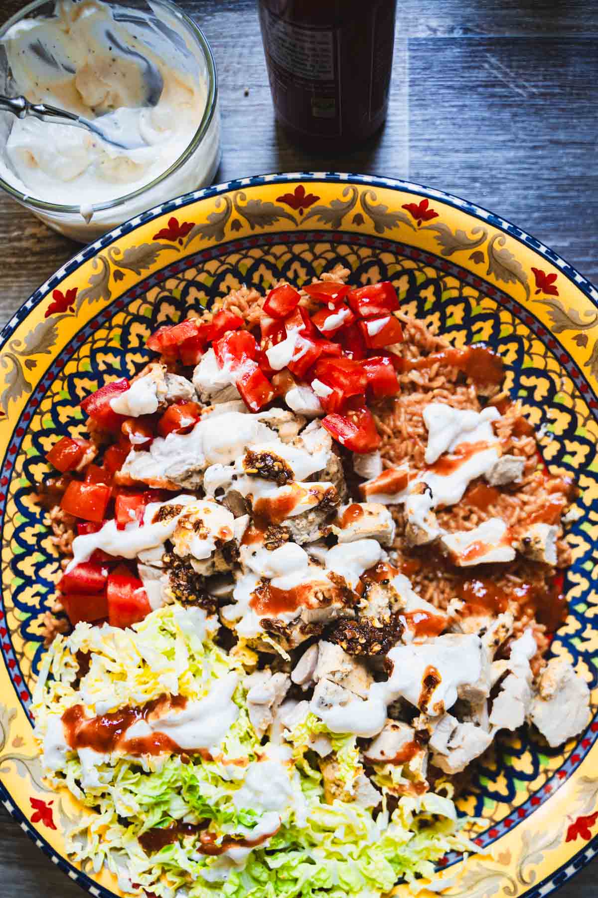 halal style chicken and yellow rice with white sauce and halal hot sauce, diced tomatoes and shredded napa cabbage in a yellow serving bowl.