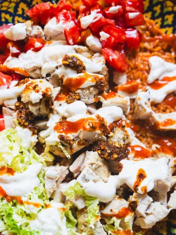 halal style chicken and yellow rice with white sauce and halal hot sauce, diced tomatoes and shredded napa cabbage in a yellow serving bowl.