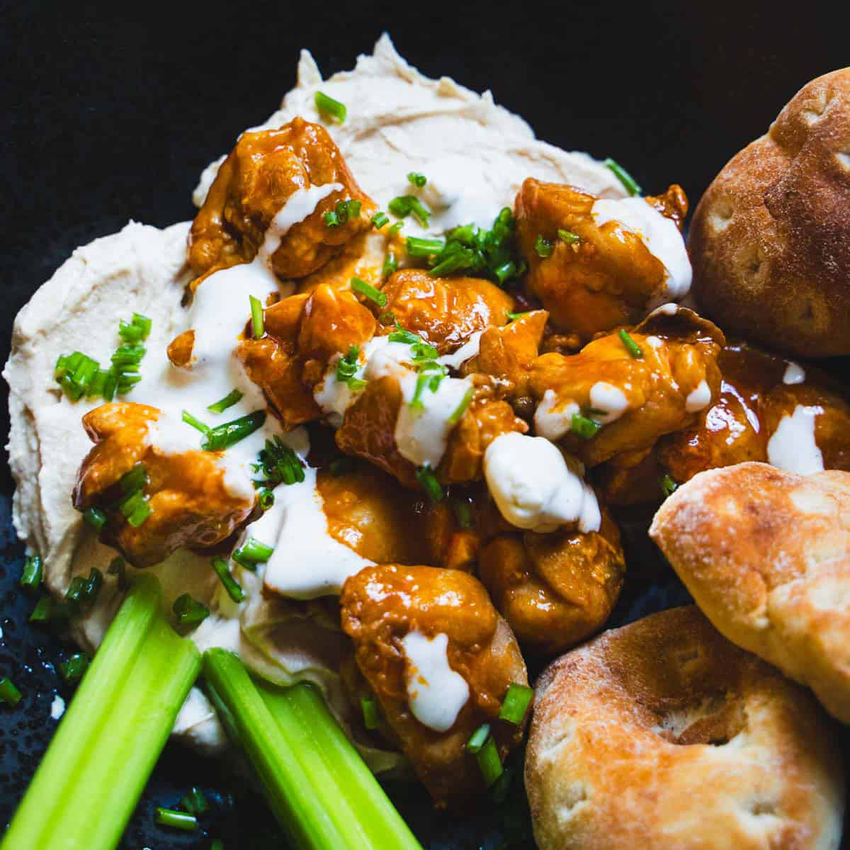 Buffalo chicken hummus bowl in a black bowl garnished with celery sticks and pita rounds next to a small bowl of blue cheese dip.
